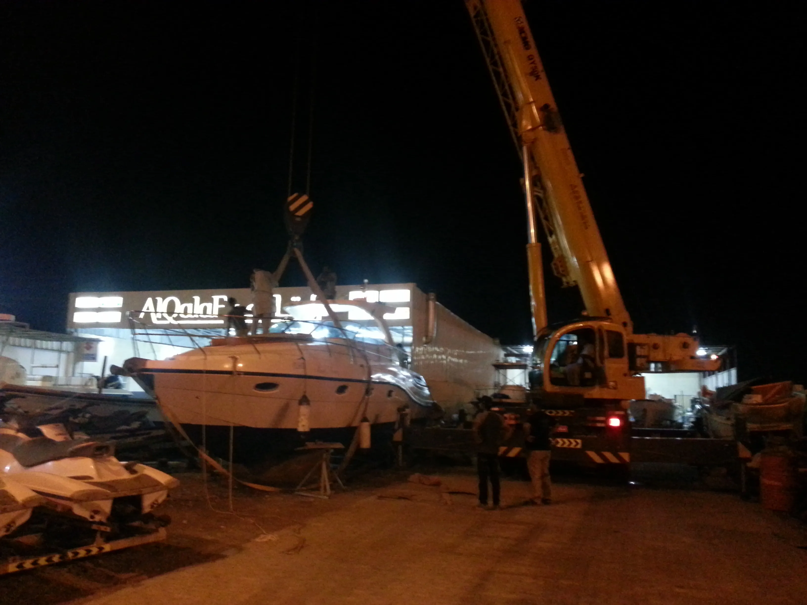 A customer's yacht being lifted by mobile crane in the workshop yard at Saham, with the AlQalaF storefront sign behind.